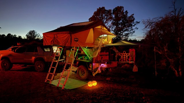 Rooftop tent camp glowing green, jack-o’-lanterns standing guard